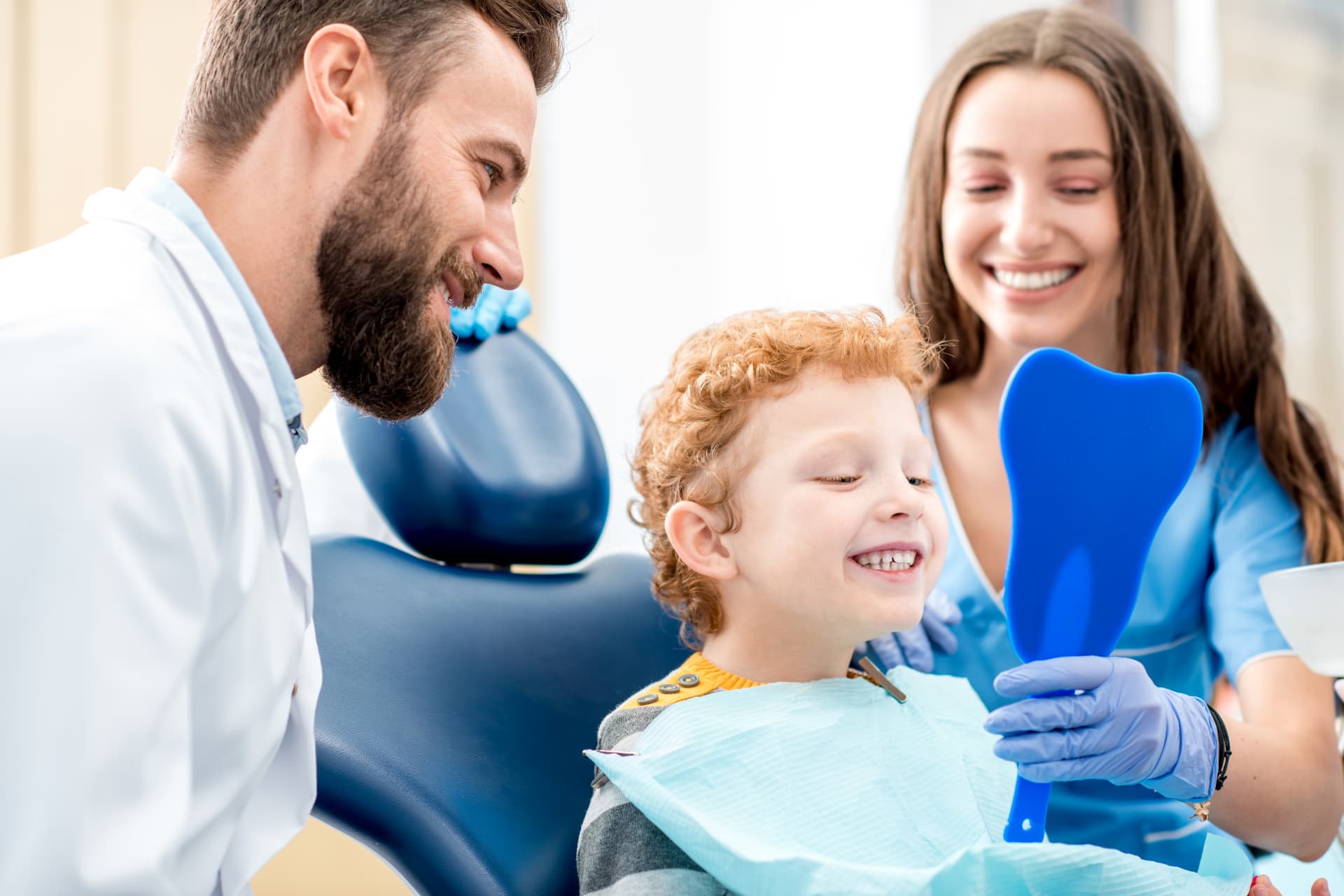 Dentist and dental assistant smiling with a young patient holding a tooth-shaped mirror